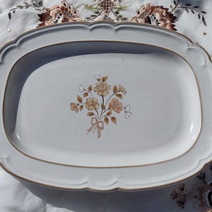 May include: A white ceramic serving platter with a scalloped edge and a floral bouquet design. The platter has a light brown trim and a rectangular shape. The floral bouquet features peach, tan, and brown flowers with a ribbon.