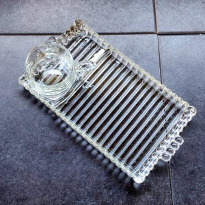 May include: A clear glass serving tray with a built-in circular bowl. The rectangular tray has a ribbed design with a beaded edge. The bowl is on the left side of the tray. The tray is designed for serving snacks or appetizers.