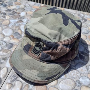 May include: Camouflage pattern military hat with a short brim. The hat features a sergeant rank insignia on the side. The colors include shades of green, brown, and black.