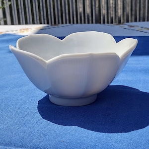 May include: A white ceramic bowl with a scalloped rim, resembling a flower. The bowl sits on a small, circular base. The bowl is on a blue surface.