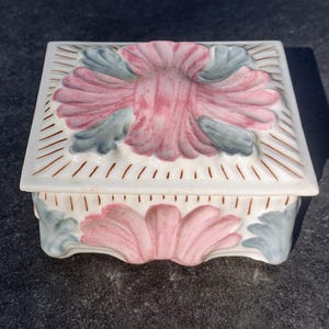 May include: A square, ceramic trinket box with a lid. The box is white with a raised floral design in pink and gray. The lid has a pink flower in the center and gray leaves. The box is likely used for jewelry or small keepsakes.