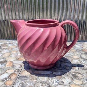 May include: A pink ceramic pitcher with a spiral design. The pitcher has a spout and a handle. It is sitting on a stone surface.