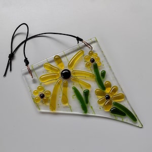 May include: A glass suncatcher with a black string hanger. The suncatcher features a yellow flower with a black center and green leaves. There are smaller yellow flowers with black centers on the suncatcher.