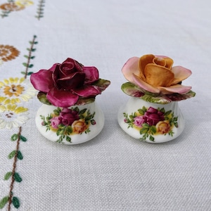 May include: Two ceramic rose figurines. One rose is deep red, the other is peach and yellow. Both are atop small, white vases decorated with rose patterns. The figurines are on a white surface with embroidered floral details.