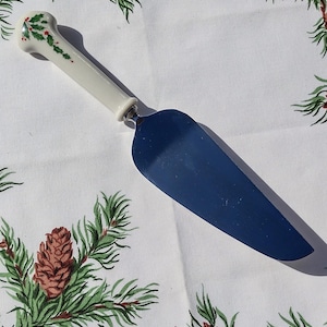 May include: A cake server with a blue blade and a white handle. The handle is decorated with green holly leaves and red berries. The server is on a white tablecloth with a green and brown pinecone pattern.