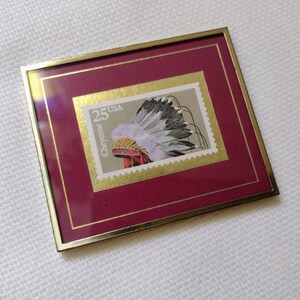 May include: Framed 25-cent USA stamp featuring a Cheyenne headdress. The stamp is set against a gold border and a deep red mat, encased in a gold-toned metal frame. The stamp's design includes a detailed illustration of the headdress.