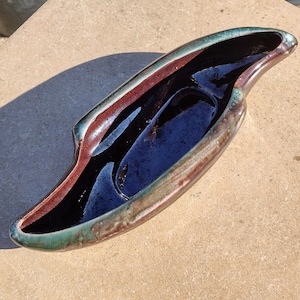 May include: A ceramic, oblong-shaped dish with a dark blue interior and a glossy finish. The rim features a gradient of colors, including brown, red, and green. The dish is likely used for decorative purposes or serving food.