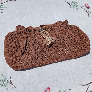 May include: A brown crocheted clutch purse with a scalloped top edge and a clear plastic clasp. The purse has an open weave design and is set against a white background with floral embroidery.