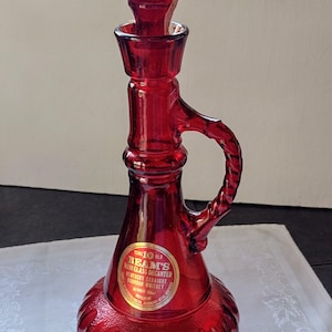 May include: A ruby red glass decanter with a handle and stopper. The decanter has a bulbous base and a label that reads "Beam's 10 Old Kentucky Straight Bourbon Whiskey". The decanter is approximately 12 inches tall.