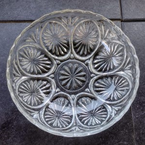 May include: Clear glass serving bowl with a decorative floral pattern. The bowl features a scalloped edge and a central starburst design, surrounded by oval-shaped floral motifs. The glass is transparent, allowing light to pass through.