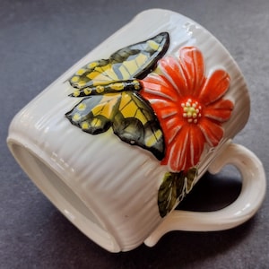 May include: White ceramic mug with a textured surface, featuring a raised design of a yellow, black, and gray butterfly and a bright red flower. The mug has a curved handle and is suitable for hot beverages.