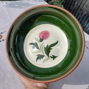 May include: A handmade ceramic bowl with a green interior and a brown rim. The center features a pink thistle flower design with green leaves on a cream background. The bowl is likely used for serving food.