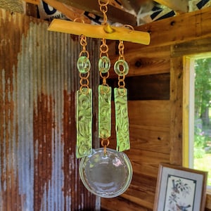 May include: A green glass wind chime with a wooden top and copper chain links. The chime has three rectangular pieces of glass with a swirling pattern and a round glass piece at the bottom.