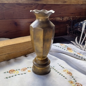 May include: A vintage brass vase with a fluted rim and a geometric, faceted body. The vase has a pedestal base and a warm, golden hue. It is displayed on a white embroidered cloth, suggesting a decorative or antique theme.