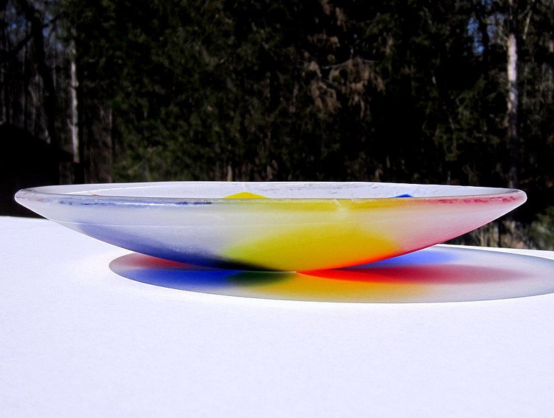 Pu&ograve; includere: Una ciotola di vetro trasparente con un design ampio e poco profondo. La ciotola presenta un gradiente di colori, che passa dal blu al giallo al rosso. La ciotola &egrave; su una superficie bianca, con uno sfondo sfocato di alberi.