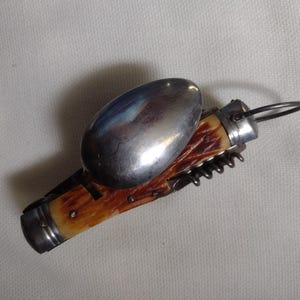 May include: A multi-tool with a spoon, corkscrew, and knife blade. The handle is a light brown color with a textured pattern. The spoon is silver and reflects light. A metal ring is attached to the end of the tool.