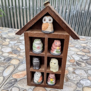May include: A wooden house-shaped display case with eight miniature animal figurines. The figurines include an owl, frog, squirrel, cat, and rabbit. The display case is brown and has a small metal loop at the top for hanging.