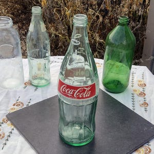 May include: A collection of vintage glass bottles, including a Coca-Cola bottle with a red label. The bottles are various shapes and sizes, with a clear and green tint. The Coca-Cola bottle is labeled "32 FL OZ (1 QT)".