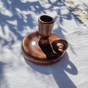 May include: A handcrafted, brown ceramic candle holder. The candle holder features a circular base with a curved handle and a cylindrical cup to hold a candle. The piece is glazed with a glossy finish, and sits on a white surface.