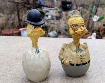 May include: Two whimsical chick figurines. One chick is wearing a black bowler hat and a white shirt with a blue tie. The other chick is wearing glasses, a tan shirt, and a gray skirt. Both are yellow and have a cracked egg base.