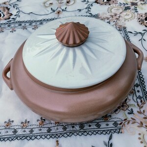 May include: A vintage, two-handled serving dish with a lid. The dish is a warm, muted brown color, with a white lid featuring a starburst design. The lid is topped with a brown, shell-shaped handle. The dish is likely ceramic.
