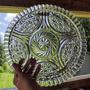 May include: A clear, vintage glass serving plate with a starburst and geometric pattern. The plate has a scalloped edge and is held up against a background of green foliage and a blue sky. The plate is round and has a decorative design.