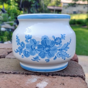 May include: A white ceramic jar with a blue floral design. The jar has a rounded shape with a blue rim and base. The floral pattern features blue flowers and leaves, adding a decorative touch to the piece.