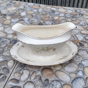 May include: A white porcelain gravy boat with a matching saucer. The boat has a curved handle and a gold trim. Floral patterns in pink and green decorate the saucer and the boat's interior.