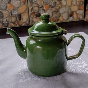 May include: A vintage green enamel teapot with a curved spout and a rounded handle. The teapot has a matching lid with a small knob. The teapot is sitting on a white tablecloth with a floral pattern.
