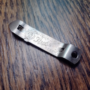 May include: A vintage metal bottle opener with the text "Stroh's Beer" and "A Quality Opener For A Quality Product". The opener is silver-colored and shows signs of wear, suggesting it is an antique or a well-used item.