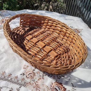 May include: A woven wicker basket with a handle. The basket is oval-shaped and features a detailed woven pattern on the base. The basket is made of light brown wicker and is sitting on a white tablecloth.