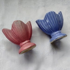 May include: Two ceramic tulip-shaped vases. One is a dusty rose color, and the other is a light blue. Both have a ribbed texture and sit on a pedestal base. The vases are likely vintage or antique decorative items.
