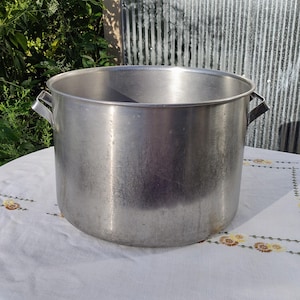 May include: A large, brushed aluminum pot with two side handles. The pot is cylindrical with a wide opening and sits on a white tablecloth with a floral pattern. The background includes greenery and a corrugated metal surface.