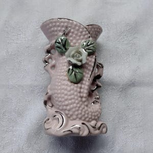 May include: A pink ceramic vase with a textured surface and a decorative rose and leaf design. The vase has a silver trim and measures approximately 6 inches tall.