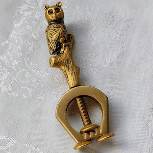 May include: A gold-colored metal corkscrew featuring an owl perched on a branch. The owl is detailed with textured feathers and sits atop a branch that forms the handle. The corkscrew mechanism is visible below the owl.