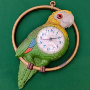 May include: A vintage parrot-shaped wall clock. The clock features a yellow and green parrot design with a gold-colored ring. The clock face has black numerals and hands. The clock is a decorative item.