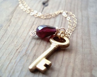May include: A gold-colored key pendant necklace with a faceted, teardrop-shaped, deep red gemstone. The necklace chain is delicate and gold, creating a vintage-inspired accessory.