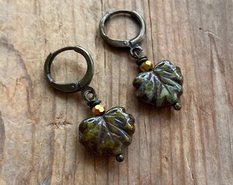 May include: A pair of antique bronze-toned earrings. Each earring features a leaf-shaped charm with green and brown hues, accented with gold-colored beads. The earrings have a lever-back closure and are displayed on a wooden surface.