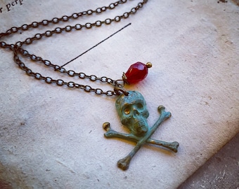 May include: A turquoise skull and crossbones pendant necklace with a red gemstone accent. The necklace is made of a brass chain.