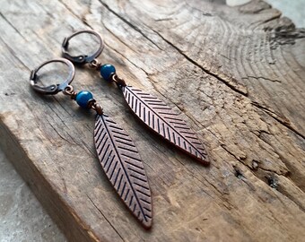 May include: A pair of copper-colored feather earrings with blue beads. The earrings have a lever back closure and are displayed on a weathered wooden surface. The feather design has detailed etched lines.