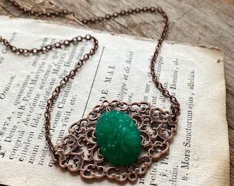 May include: A vintage-style necklace featuring a green oval pendant with a carved design, set within an ornate copper-colored filigree frame. The necklace hangs on a delicate chain, resting on a vintage book page.