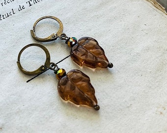 May include: A pair of dangle earrings featuring brown leaf-shaped glass pendants. The earrings have bronze-colored lever-back closures and are accented with iridescent beads. The earrings are set against a backdrop of aged paper.