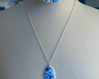 May include: A necklace and earring set featuring a blue and white porcelain design. The necklace has a silver chain with an oval pendant. The earrings are teardrop-shaped. The jewelry is displayed on a mannequin.