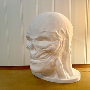May include: A white, sculpted bust of a creature with a textured, weathered appearance. The sculpture features a detailed face with prominent features and a rounded head. The bust sits on a circular base, and the overall design suggests a fantasy or horror theme.