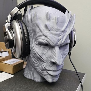 May include: A gray Night King head statue with headphones. The statue has a detailed, textured surface and is wearing black and silver headphones. The headphones are connected to a black cable. The statue is on a dark gray surface.
