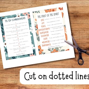 May include: Two printed pages with floral borders display "The Ten Commandments" and "The Fruit of the Spirit" texts. Black scissors are positioned near the pages. A white banner at the bottom reads "Cut on dotted lines". The background is a wooden surface.