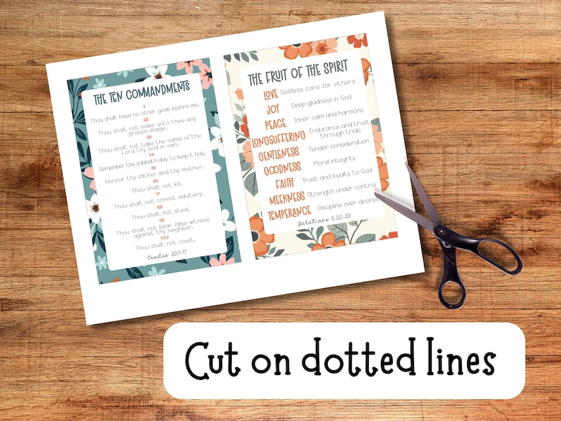 May include: Two printed pages with floral borders display "The Ten Commandments" and "The Fruit of the Spirit" texts. Black scissors are positioned near the pages. A white banner at the bottom reads "Cut on dotted lines". The background is a wooden surface.