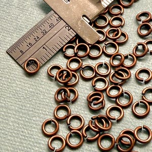 May include: Close-up of copper-colored jump rings scattered on a light green surface. A metal ruler with measurements in inches is partially visible, indicating the size of the rings. The rings are open and ready for use in jewelry making or crafts.