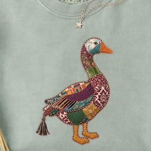 Op de afbeelding: Een lichtgroen Comfort Colors T-shirt met een geborduurd ganzenontwerp. De gans is gemaakt van kleurrijke patchworkstof met een oranje snavel en poten. Een zilveren ketting met een sterhanger ligt op het shirt.