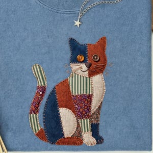 May include: Blue jean colored Comfort Colors t-shirt with a patchwork cat design. The cat is made of various fabrics in shades of blue, orange, and green. A silver necklace with a star pendant and a brown hat are also visible.